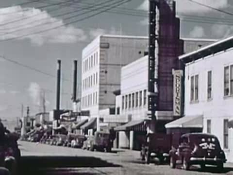 A town street with parked vintage cars and industrial brick buildings with tall chimneys.