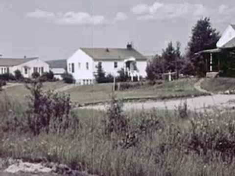 A cluster of white houses sits on a grassy hill with trees and a clear sky.