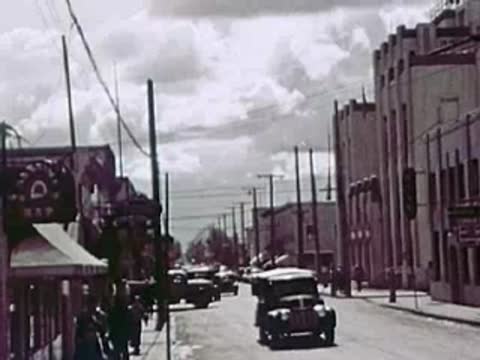 A town street with vintage vehicles driving past storefronts and overhead utility lines.