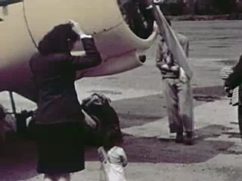 A woman and a young child stand near the engine of a parked airplane while a crew member works nearby.
