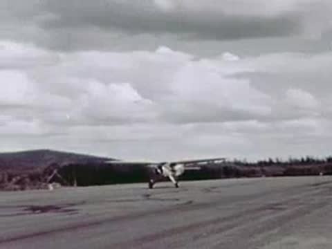 A small airplane taxis across a wide gravel airfield under a partly cloudy sky.