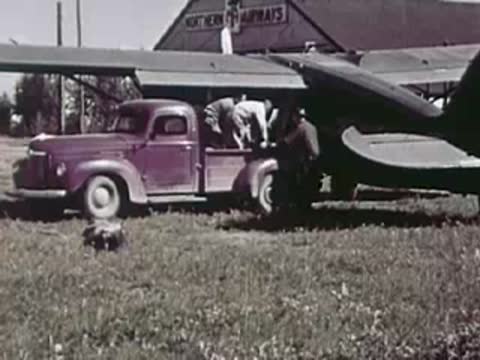 Men loading cargo into a vintage pickup truck parked beside an airplane