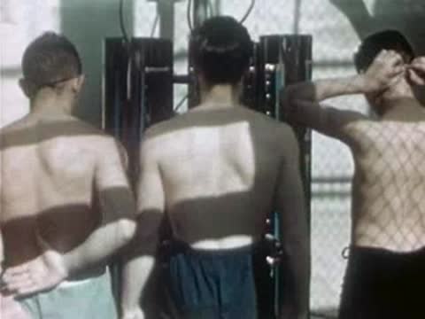 Men stand shirtless in front of vertical ultraviolet therapy lamps in a clinic room.