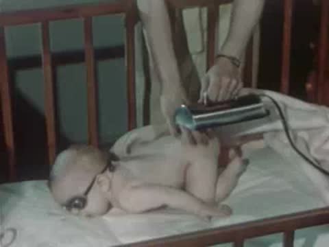 A person uses a portable ultraviolet therapy lamp on the back of an infant lying on a bed.