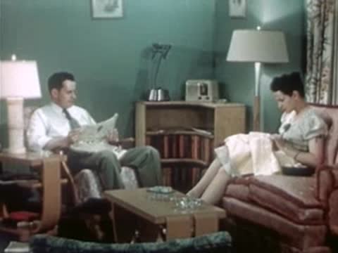 A man reads a newspaper and a woman sews in a brightly lit living room.