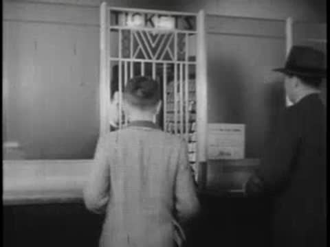 A young boy in a jacket approaches a ticket window with a man standing nearby
