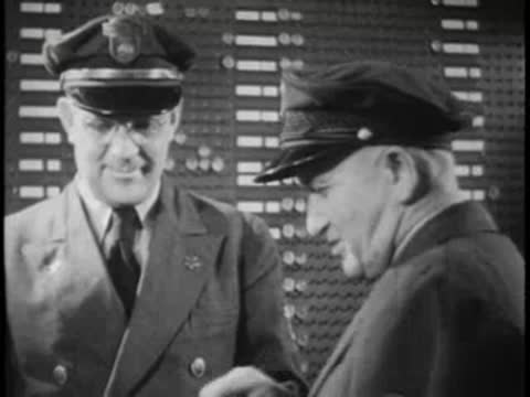 Two bus drivers talking, wearing uniforms and caps, office background.