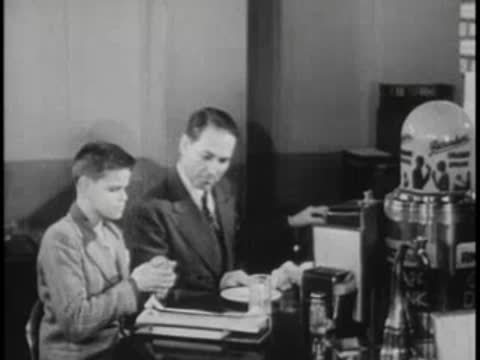 A man in a suit talking to a boy seated at a desk or counter.