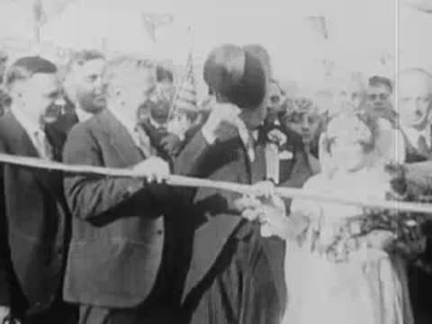 Officials and a woman in a crown cut a large ceremonial ribbon outdoors.