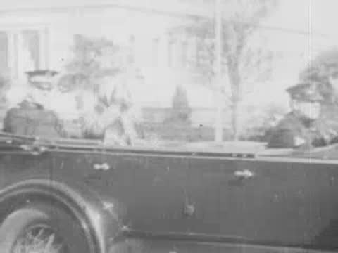 Vintage open-top car drives along a street lined with spectators and trees.