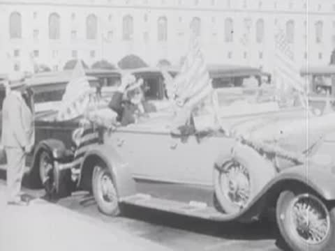 Vintage open-top car with flags drives slowly past onlookers on a sunny day.