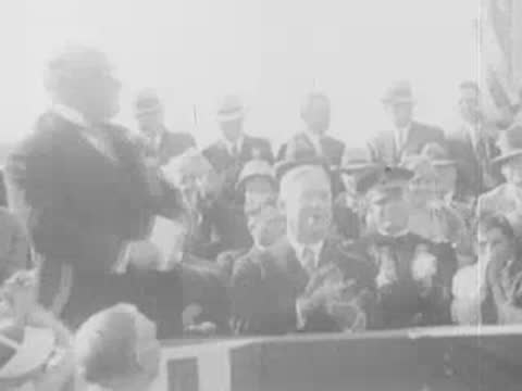 Crowd watches as officials speak at a microphone on a flag-draped stage.