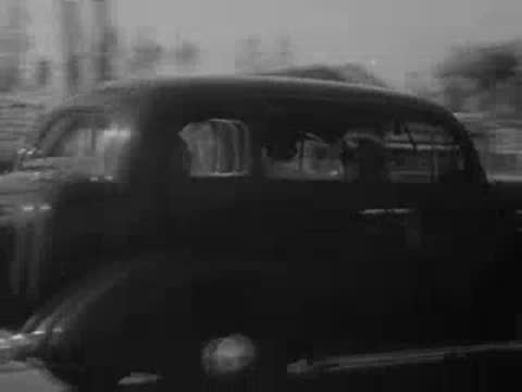 A black sedan drives along a street lined with palm trees.