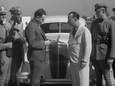 Men in uniform and suits shake hands outdoors near parked cars.