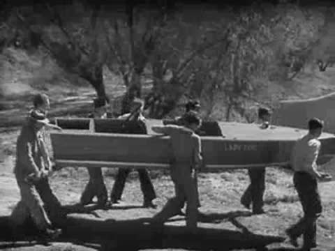 Men lift a boat off a car roof rack by a riverbank.