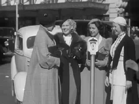 Four women stand chatting on a city sidewalk by a parking meter.