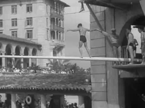 men walk along a long horizontal pole above a swimming pool