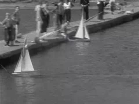 Spectators watch as model sailboats are launched from a concrete dock into water.