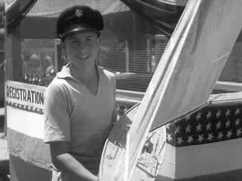 A young man in a sailor hat holds a model sailboat on a deck.