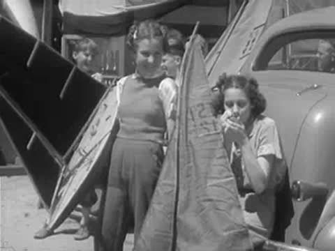 Two children stand near a car holding a model sailboat.