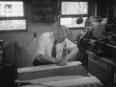 A man meticulously crafts wooden parts for a model sailboat in a workshop.