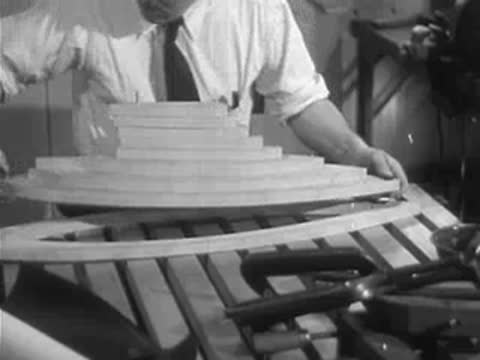 A man assembles layered wooden hull pieces of a model sailboat on a workbench.