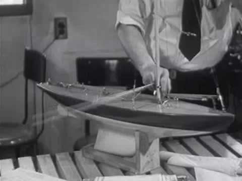 A man carefully rigs the sails and rigging on a completed model sailboat.