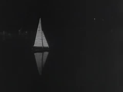 A lone model sailboat glides across dark water illuminated by a spotlight at night.