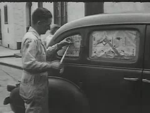 A man in coveralls applying tape to the side window of a car.