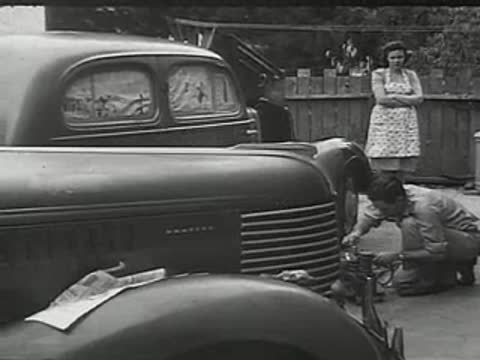 A man working on equipment in front of a car with a woman watching.