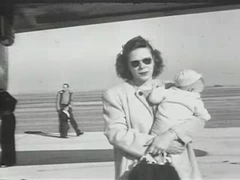 A woman holding a baby on an airfield with people and aircraft nearby.