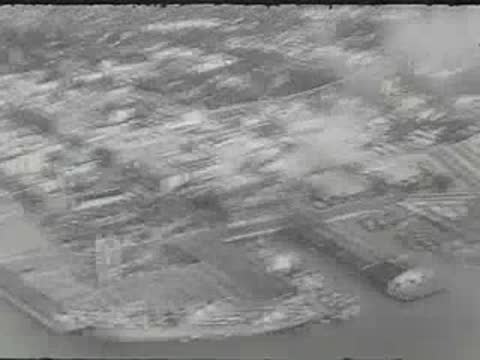 An aerial tracking shot of a port city with docked ships and smoke.