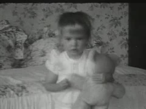 A toddler in a white dress sits on a bed playing with a doll.