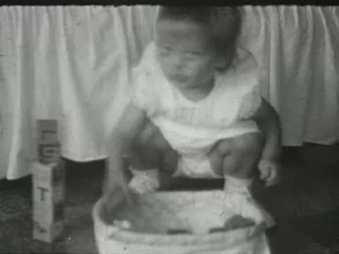 A toddler in a white dress squats in front of a white laundry basket indoors.