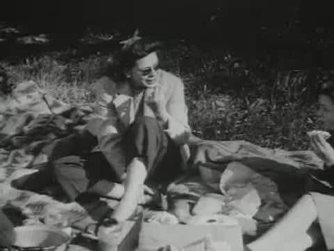 A woman sits on a picnic blanket outdoors wearing sunglasses and eating.