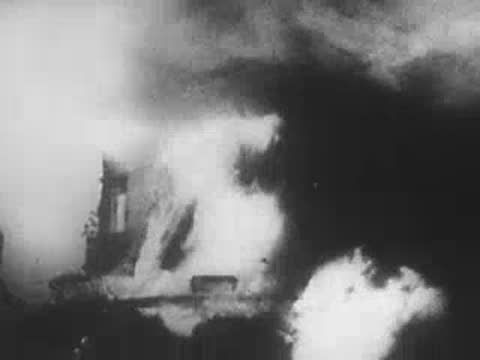 View looking down from a ship deck toward an explosion and smoking town.