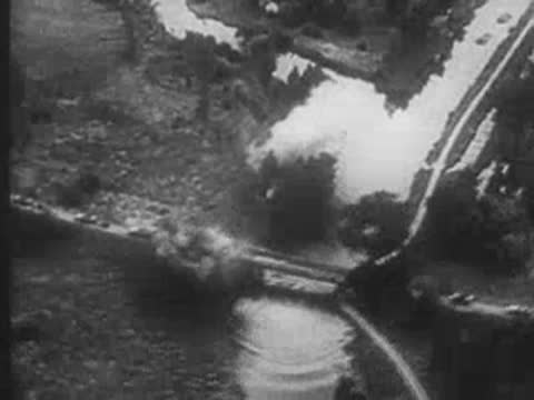military vehicles crossing a bridge with explosions and smoke nearby