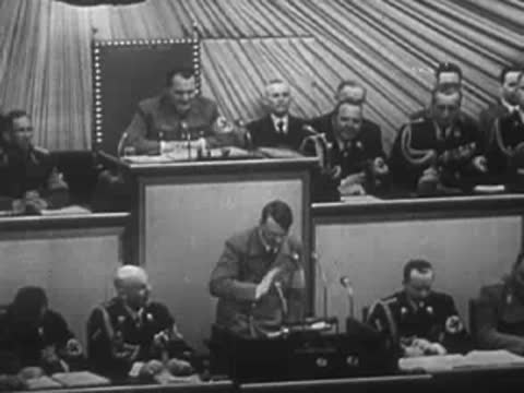 Adolf Hitler speaks at a podium in front of seated officials.