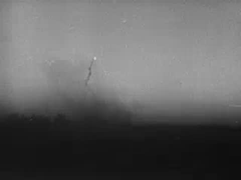 Dark landscape at dusk or night with distant muzzle flashes and fire sparks.
