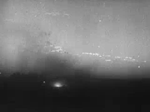 Nighttime landscape with faint lights and smoke rising from the terrain.