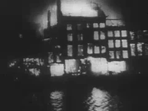 Silhouetted burning buildings along a waterfront reflecting in the water at night, WWII
