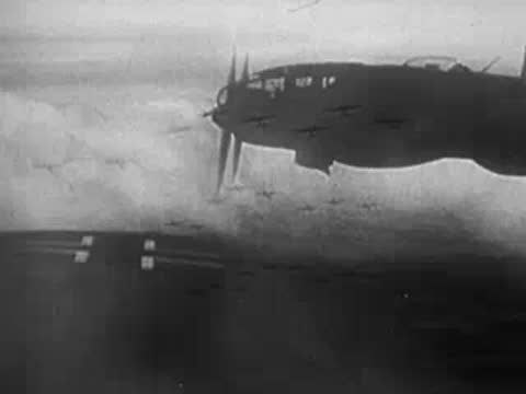Military aircraft in flight with clouds and ground visible below.