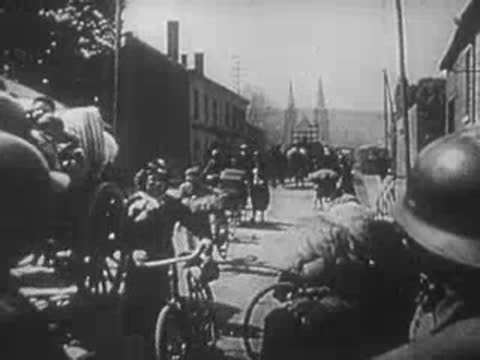 Horse-drawn carts and cyclists moving along a crowded village street.