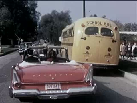 Following shot behind a red convertible driving past a yellow school bus.
