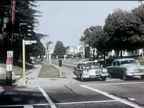 cars driving down a suburban street past lush green trees, 1950s