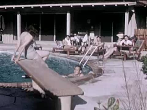 Woman sits on a diving board by a swimming pool with people relaxing nearby.