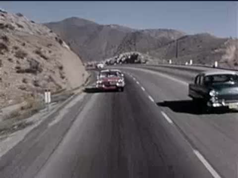 cars driving along a sunny highway next to arid rocky hills, 1950s