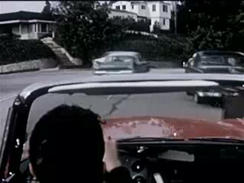 driver in a convertible navigating a suburban street with parked cars, 1950s