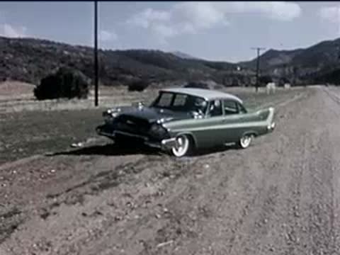 A green vintage car driving along a dirt road in a rural area.