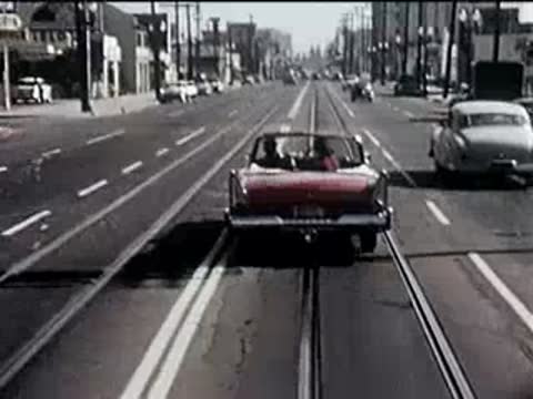 Rear view of a red convertible driving down a busy city street with tram tracks.